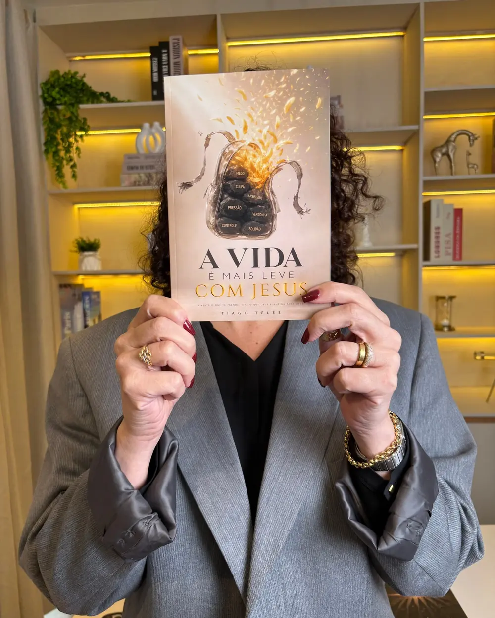 Leitora segurando o livro A Vida É Mais Leve com Jesus diante de uma estante iluminada